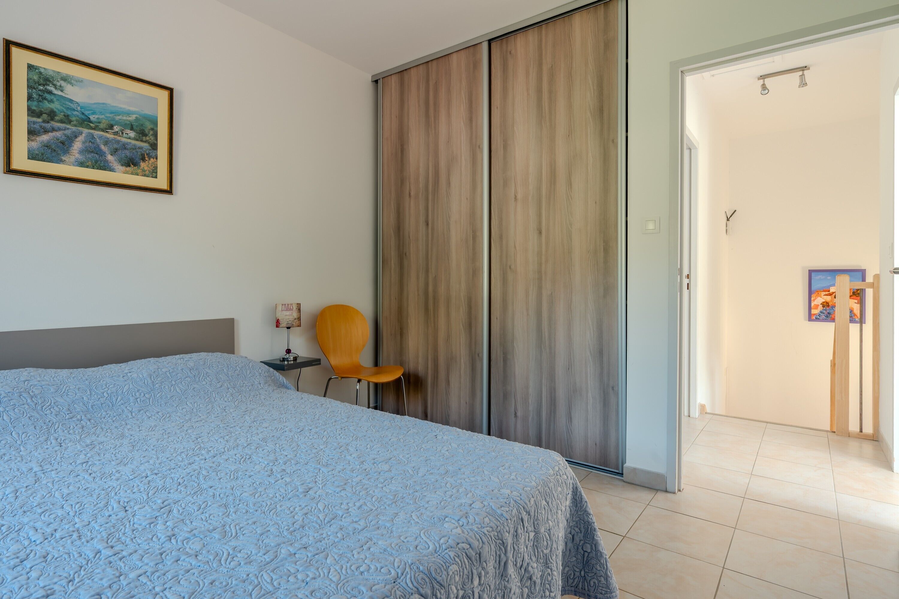 A bedroom with a bed, a chair, a wardrobe, and a door leading to another room.