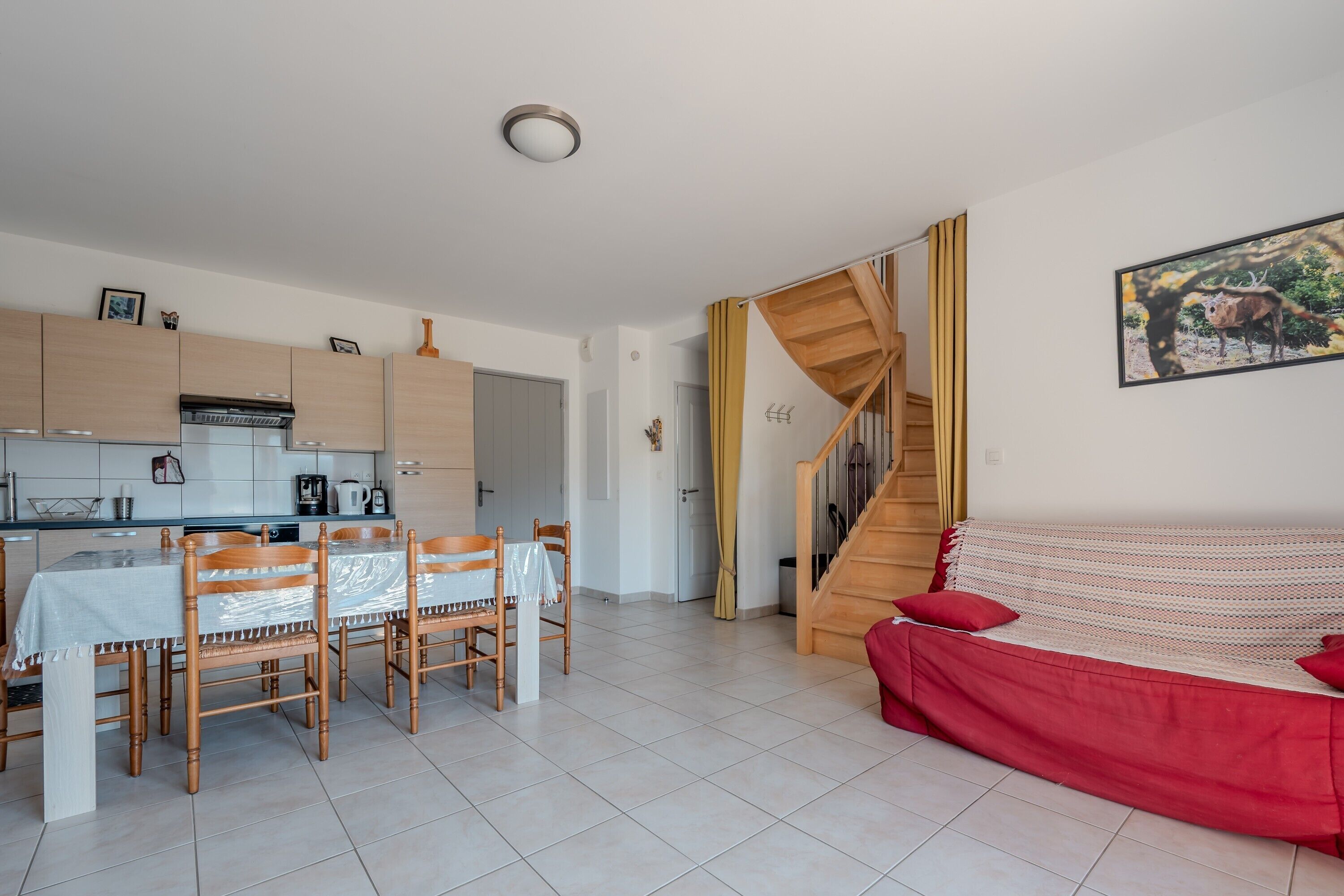 Cozinha moderna com armários de madeira, área de refeições com cadeiras e mesa, escada para outro nível e um sofá vermelho na sala de estar.