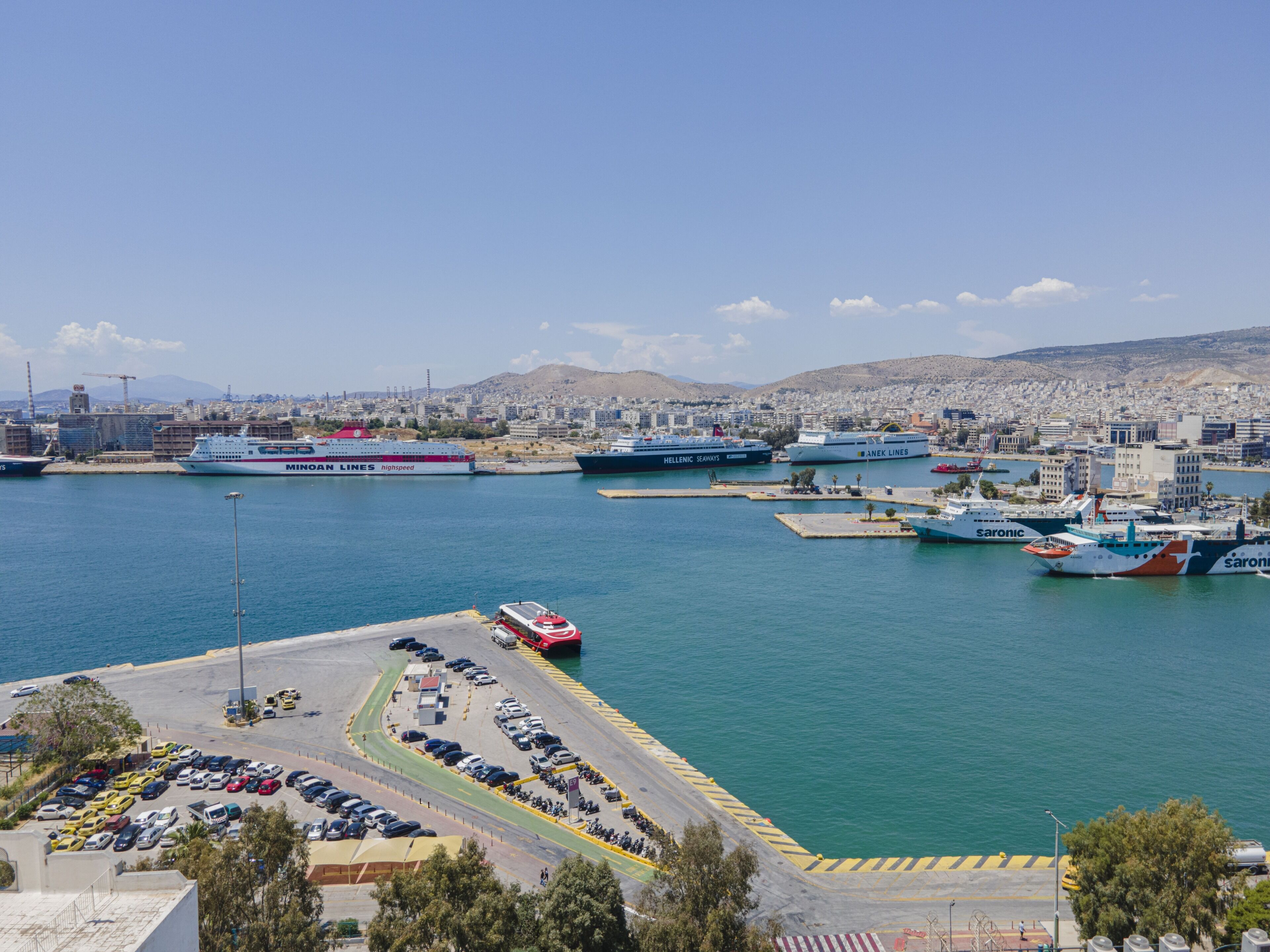 Egy kikötő, ahol több hajó is horgonyoz, egy parkoló és a háttérben egy városi panoráma látható.