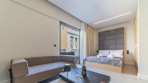 A modern hotel room with a sofa, a bed, a small table with a bottle and glasses, and a view of the city through a window.