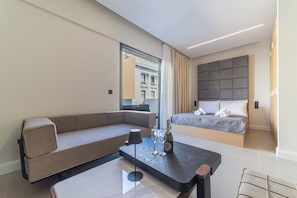 A modern hotel room with a sofa, a bed, a small table with a bottle and glasses, and a view of the city through a window. - AKVA Property - The Piraeus Collection (Pireas)