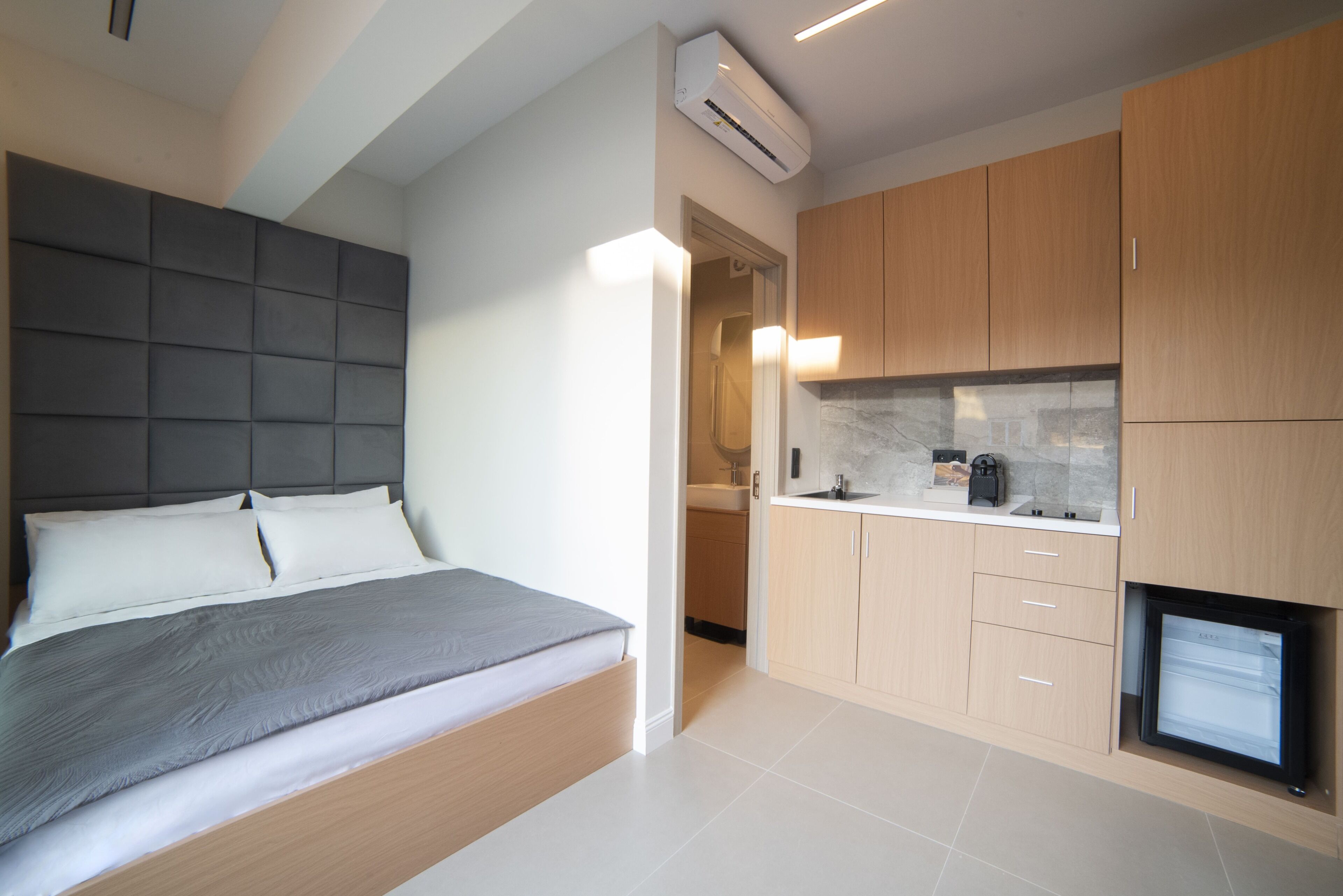 A modern bedroom with a bed, a built-in headboard, and a small kitchenette with a refrigerator and sink.