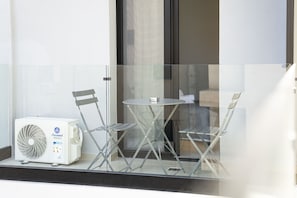 An air conditioning unit, two foldable chairs, and a small table on a balcony with a glass railing.