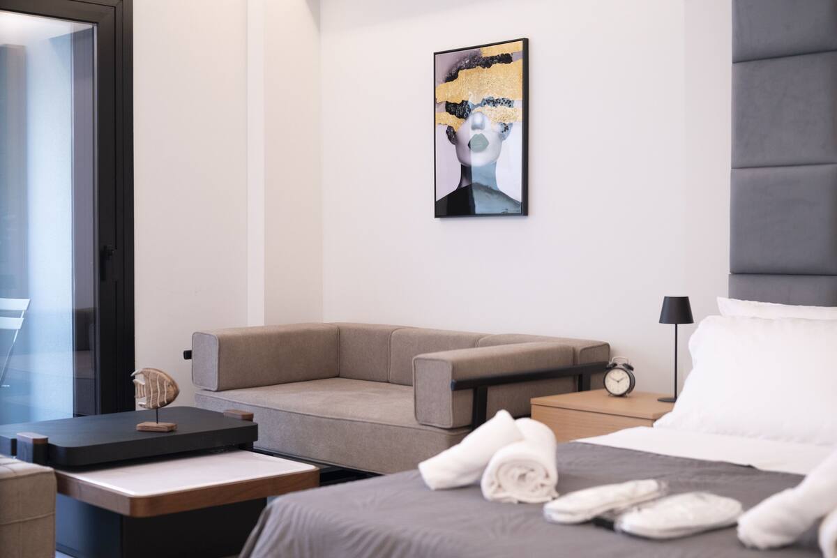 Moderni hotellihuone, jossa on sänky, sohva, yöpöytä, lamppu ja taulu seinällä.