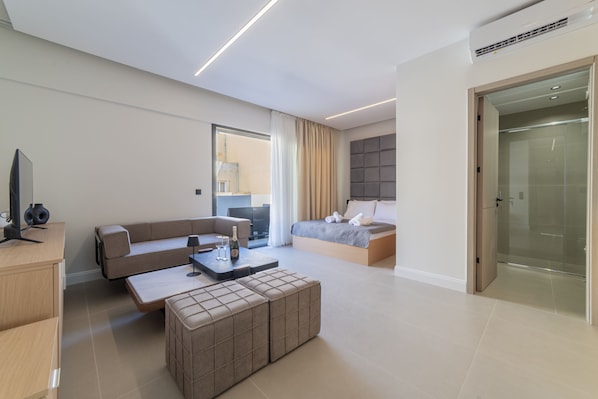 A modern hotel room with a sofa, a bed, a desk, and a glass shower enclosure. - AKVA Property - The Piraeus Collection (Pireas)