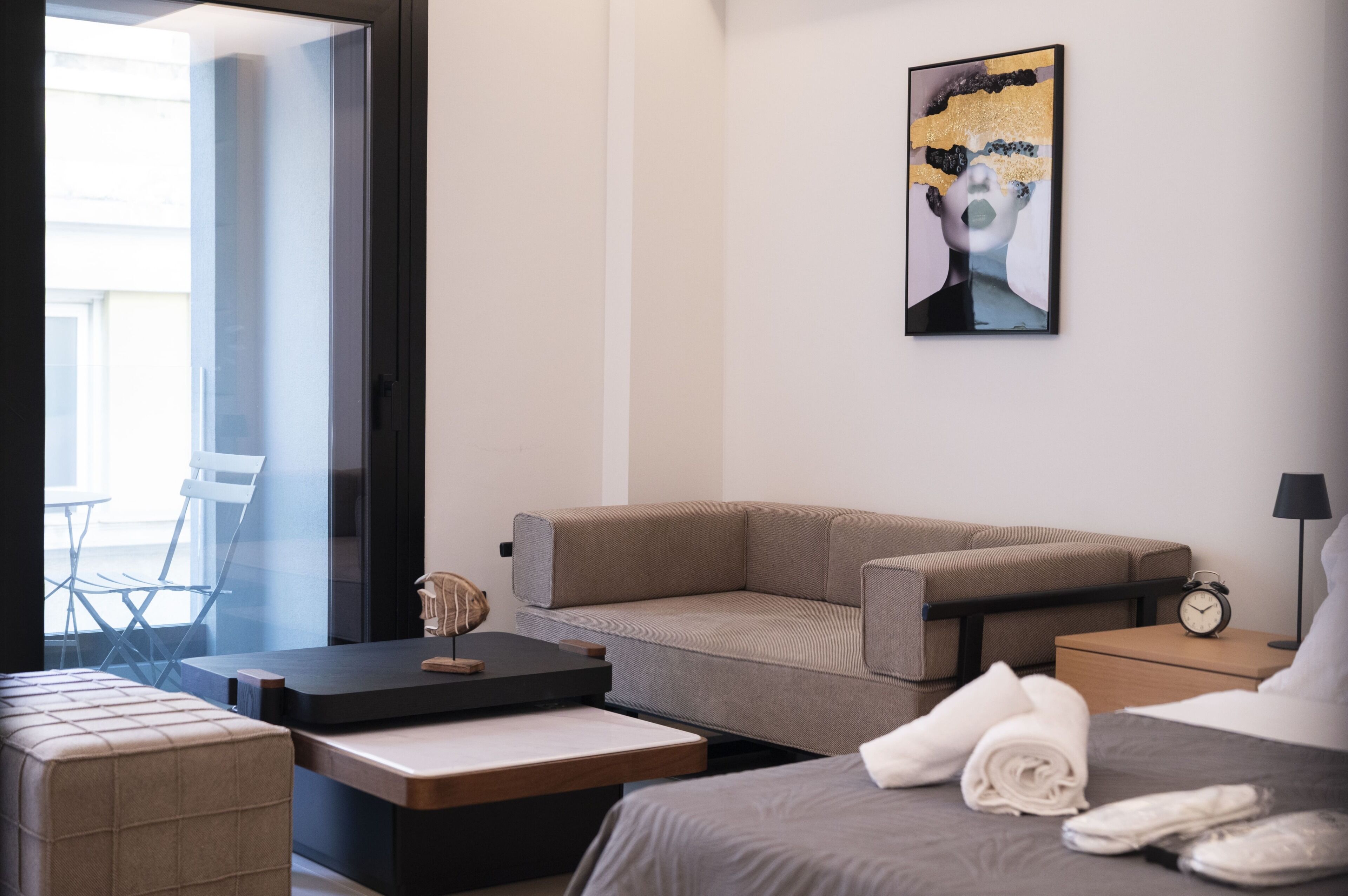 Een moderne hotelkamer met een bank, een klein tafeltje, een bed met netjes opgevouwen handdoeken, een wandklok en een schilderij.