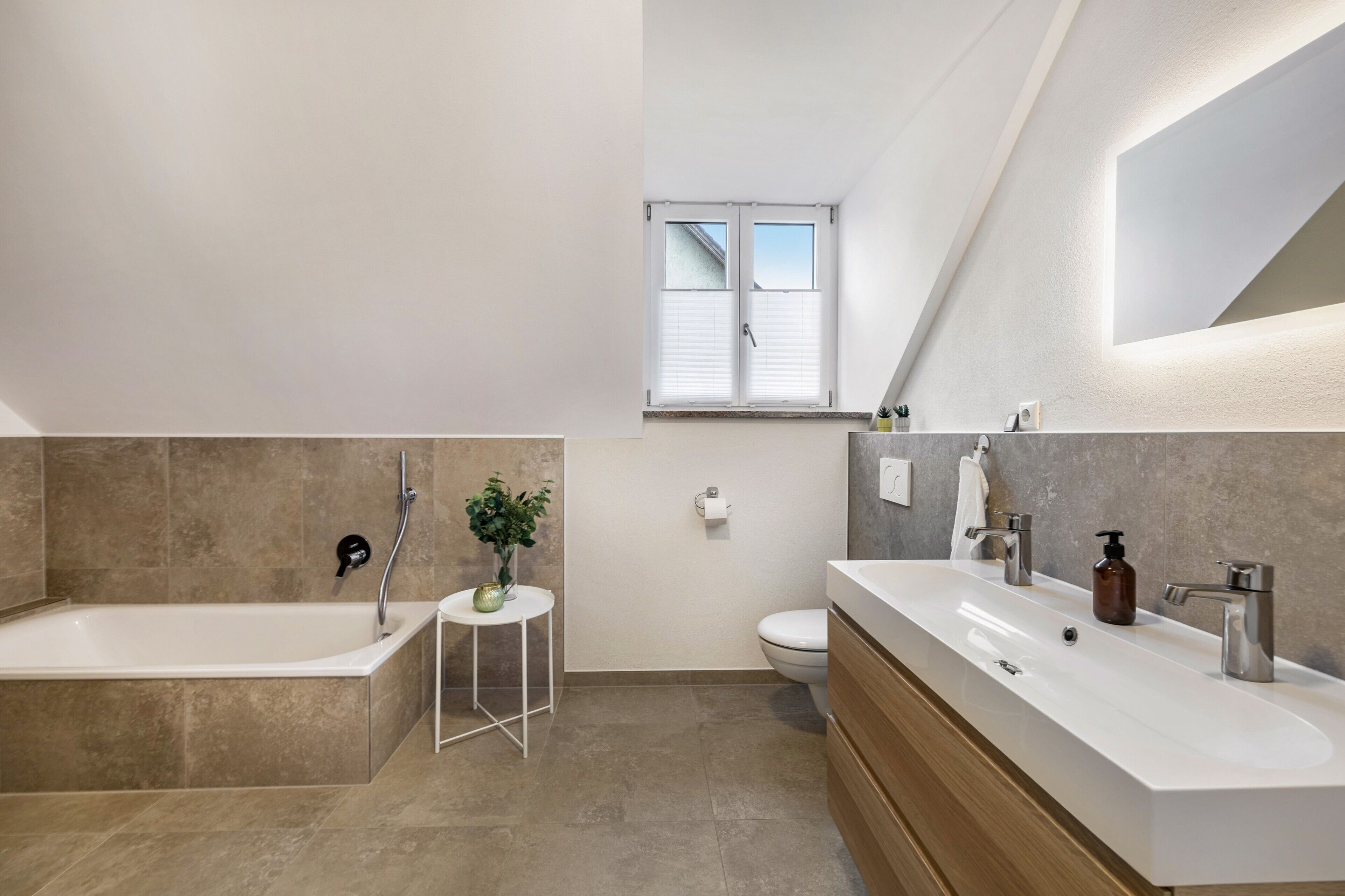 Ein modernes Badezimmer mit einer Badewanne, einem Waschbecken und einer Toilette. Über der Toilette befindet sich ein kleines Tischchen mit einer Pflanze und ein Fenster.