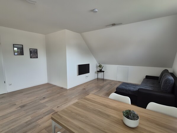 Une pièce moderne et bien éclairée, dotée d’un plancher en bois, d’une table à manger, d’un canapé et d’un téléviseur fixé au mur.