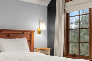 A bedroom with a wooden headboard, a bedside table with a lamp, and a window with a view of trees. - The Staghaus | Exclusive Tamarack Perks (Donnelly)
