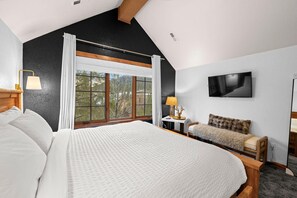 Ein Schlafzimmer mit einem großen Bett, einem Fernseher, einem Schreibtisch mit Lampe und Blick auf Bäume durch ein Fenster.