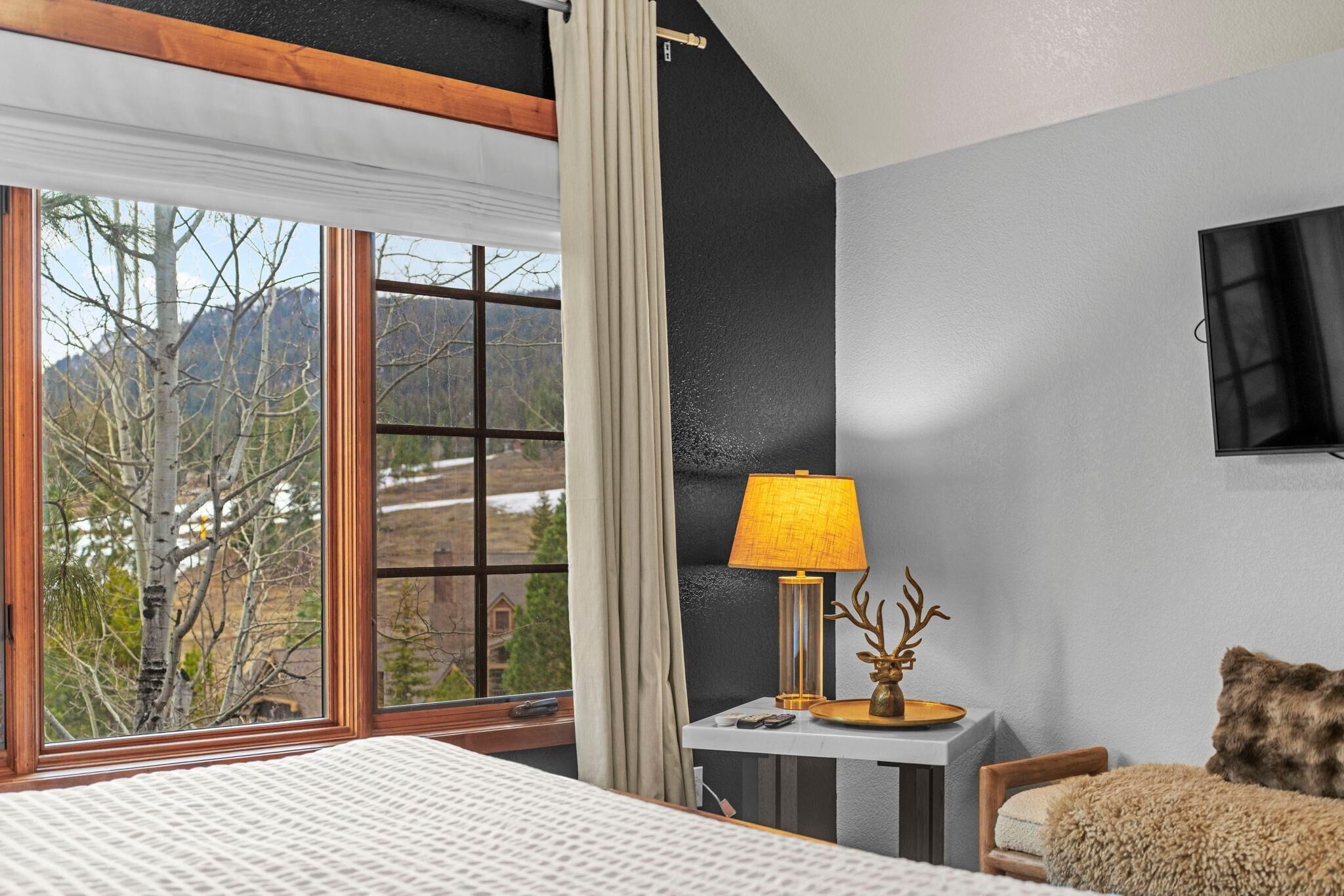 A hotel room with a bed, a desk with a lamp, a chair, a TV, and a window with a view of trees and mountains.