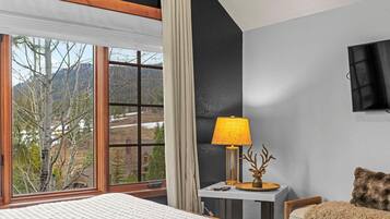A hotel room with a bed, a desk with a lamp, a chair, a TV, and a window with a view of trees and mountains.
