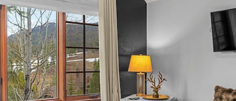 Une chambre d’hôtel avec un lit, un bureau avec une lampe, une chaise, une télévision et une fenêtre donnant sur des arbres et des montagnes.