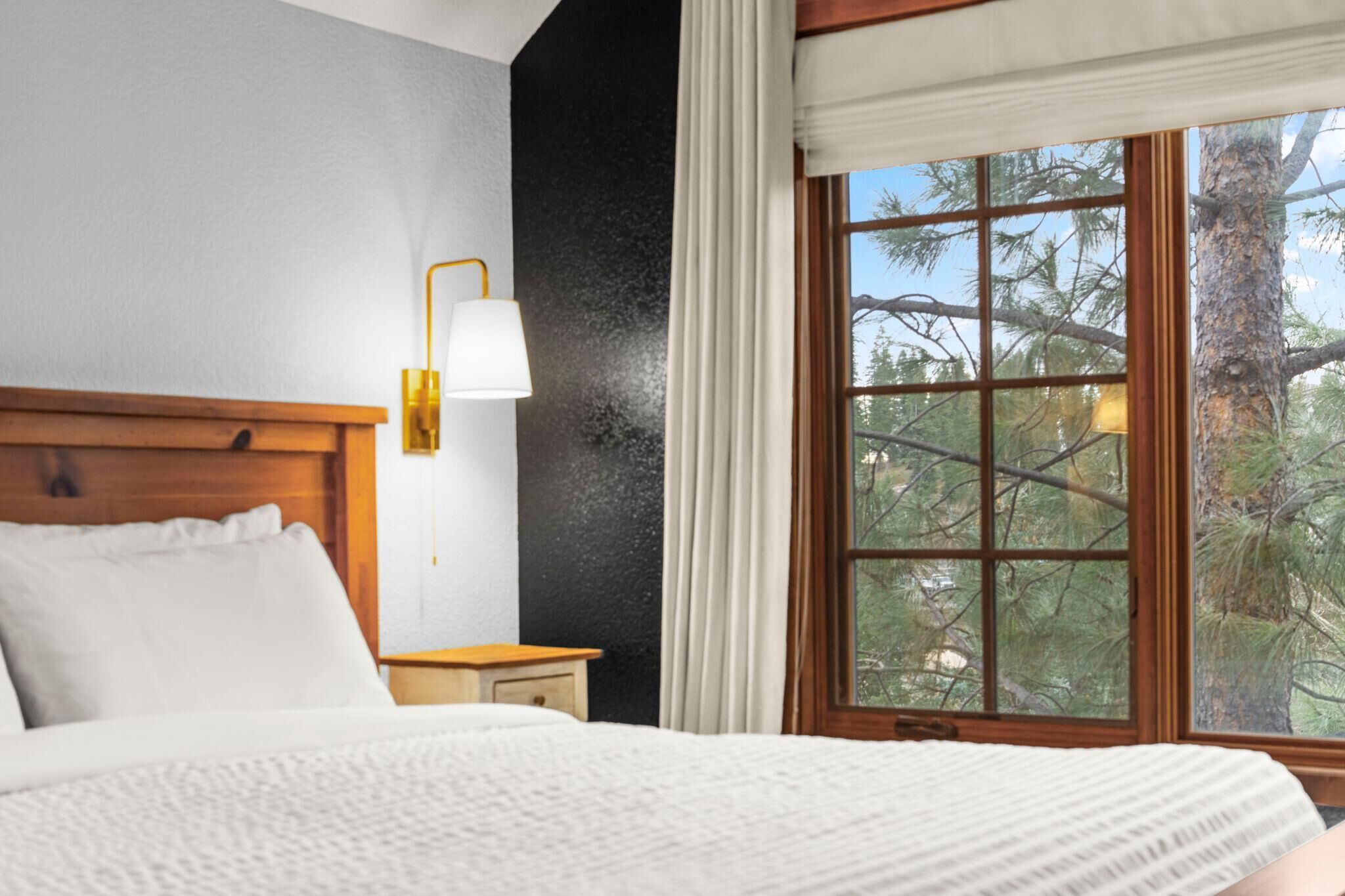 A bedroom with a wooden headboard, a bedside table, a lamp, and a window with a view of trees.