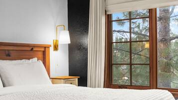 A bedroom with a wooden headboard, a bedside table, a lamp, and a window with a view of trees.