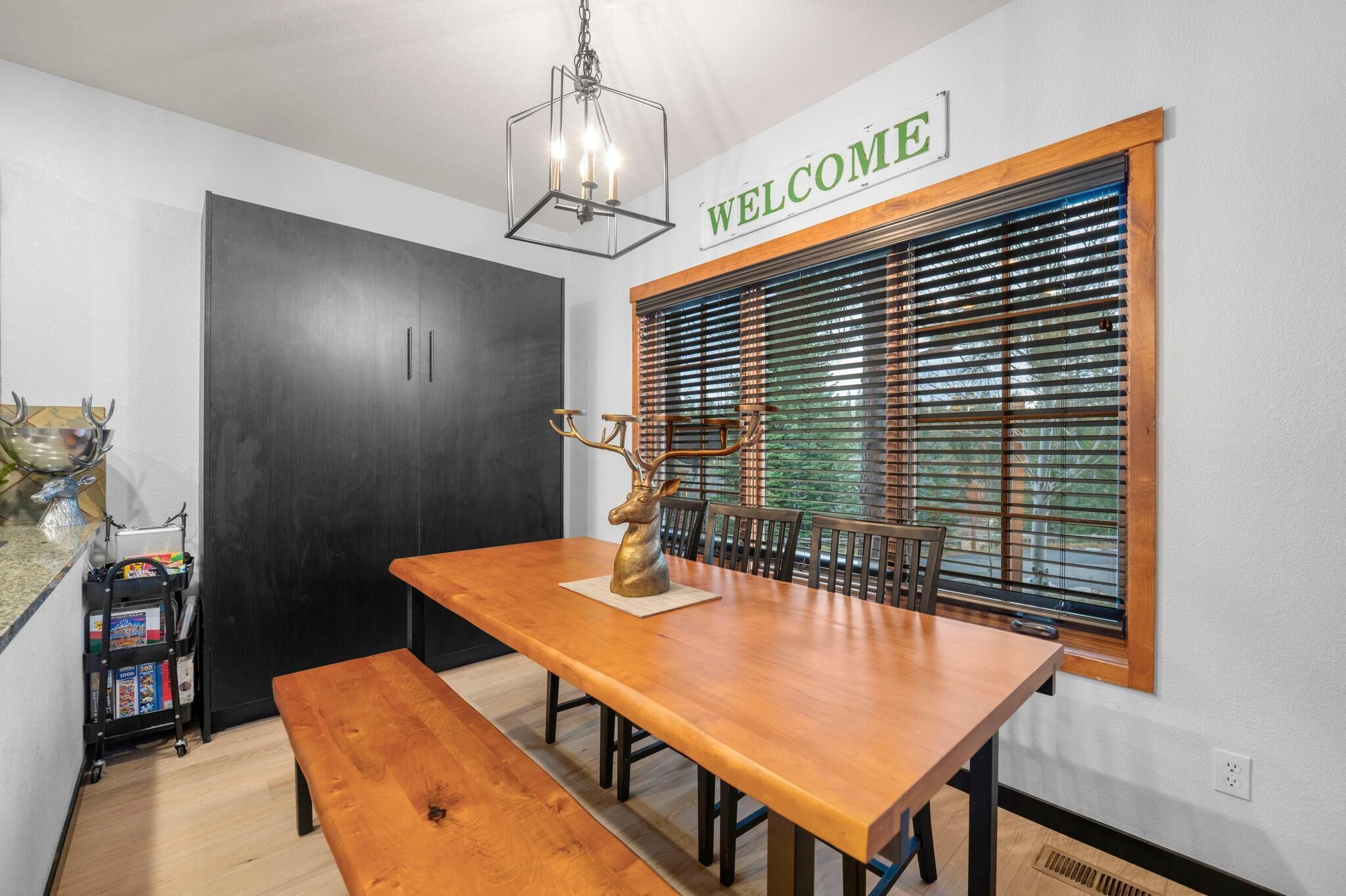 A dining area with a wooden table, benches, a deer sculpture, a 'WELCOME' sign, and a window with blinds.