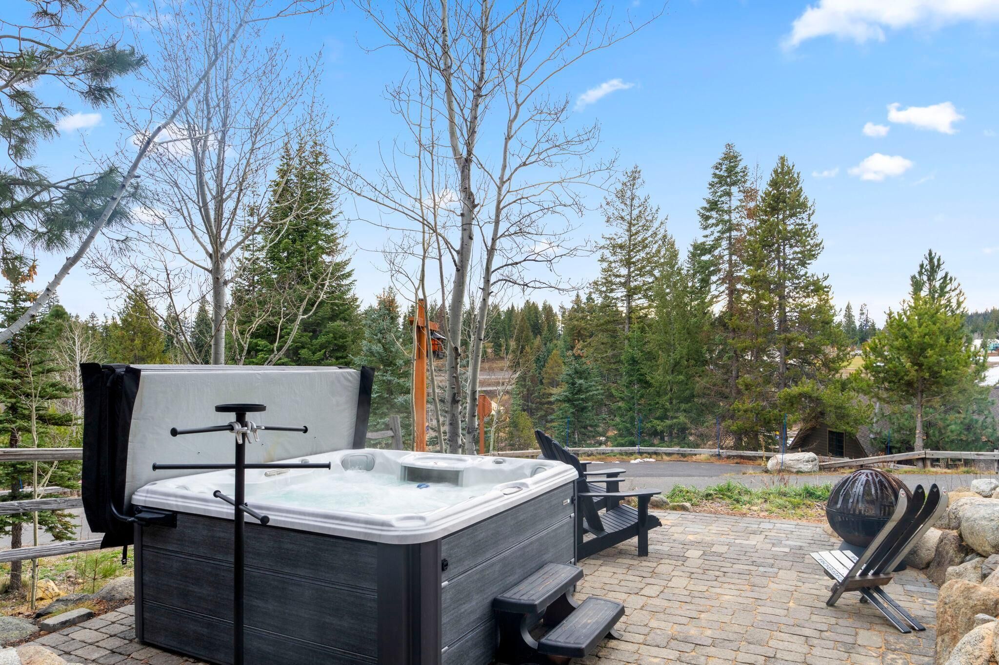 A hot tub with a wooden bench and a grill, surrounded by trees and a paved area.