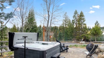 A hot tub with a wooden bench and a grill, surrounded by trees and a paved area.