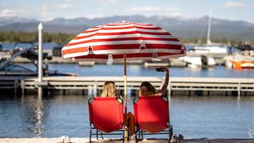 Two individuals seated under a striped umbrella at a waterfront location with boats and a dock in the background.