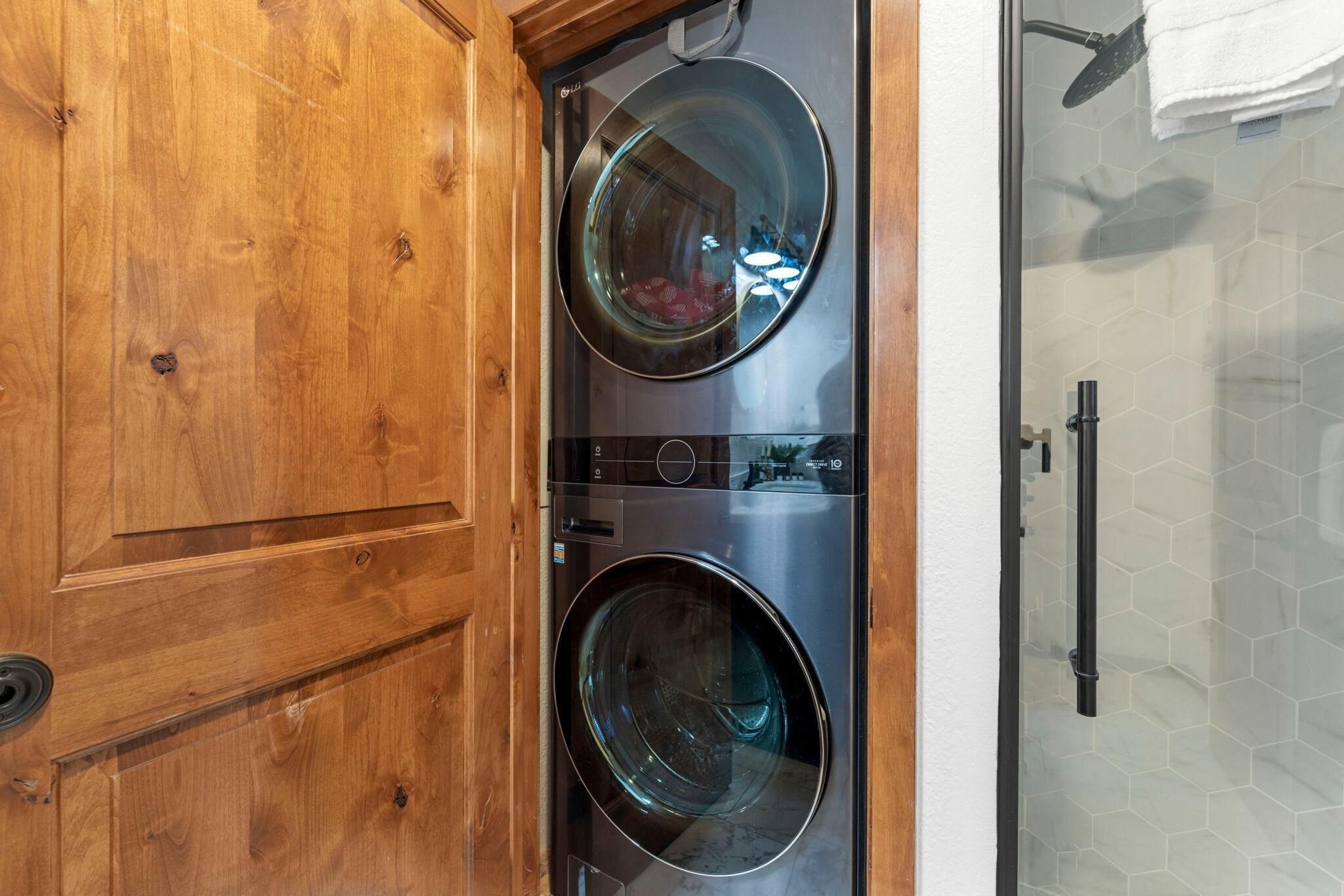 A laundry room with two front-loading washing machines, a wooden door, and a glass shower enclosure.