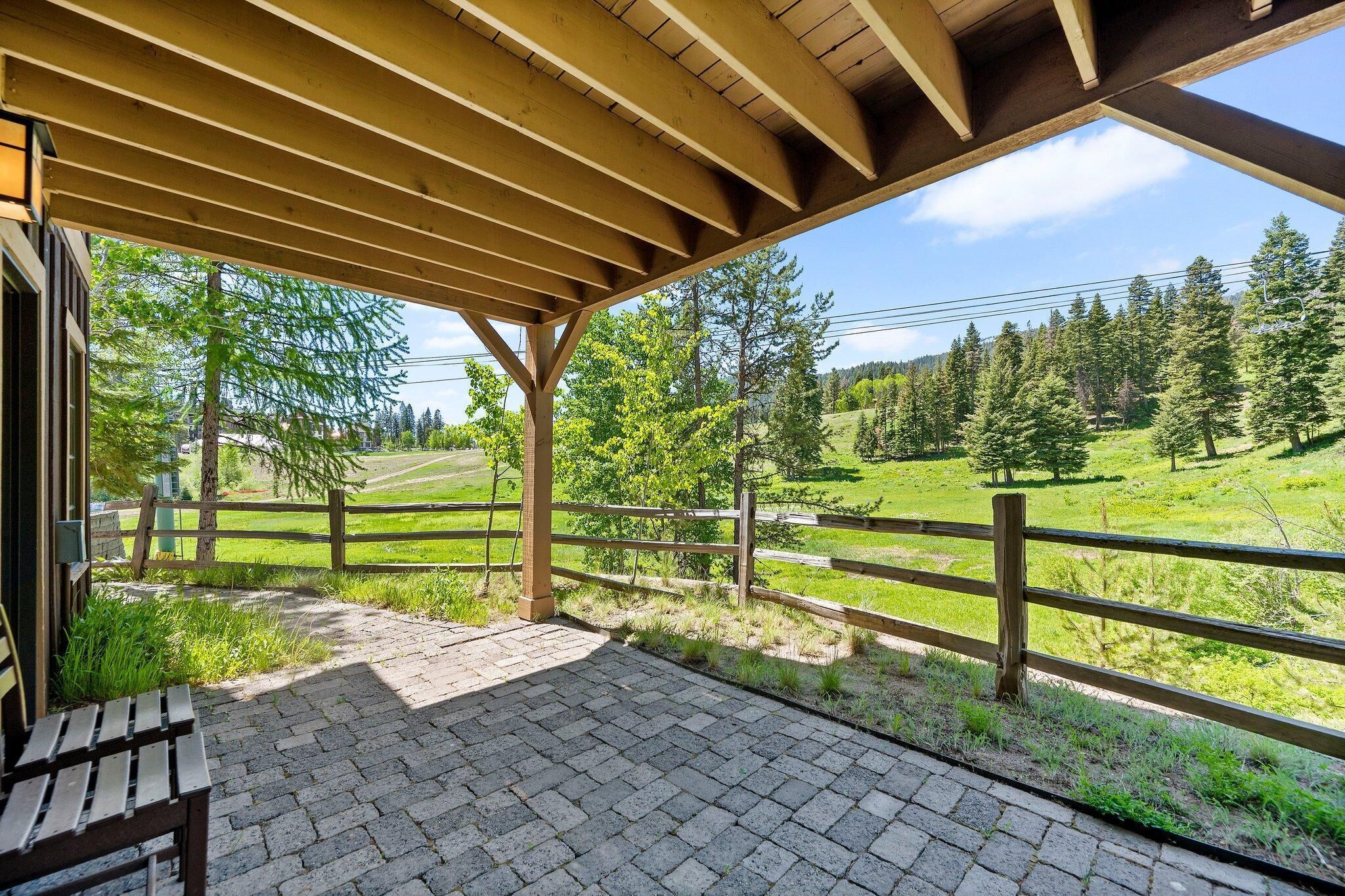 A covered outdoor area with a wooden bench and a view of a green landscape with trees and a fence.