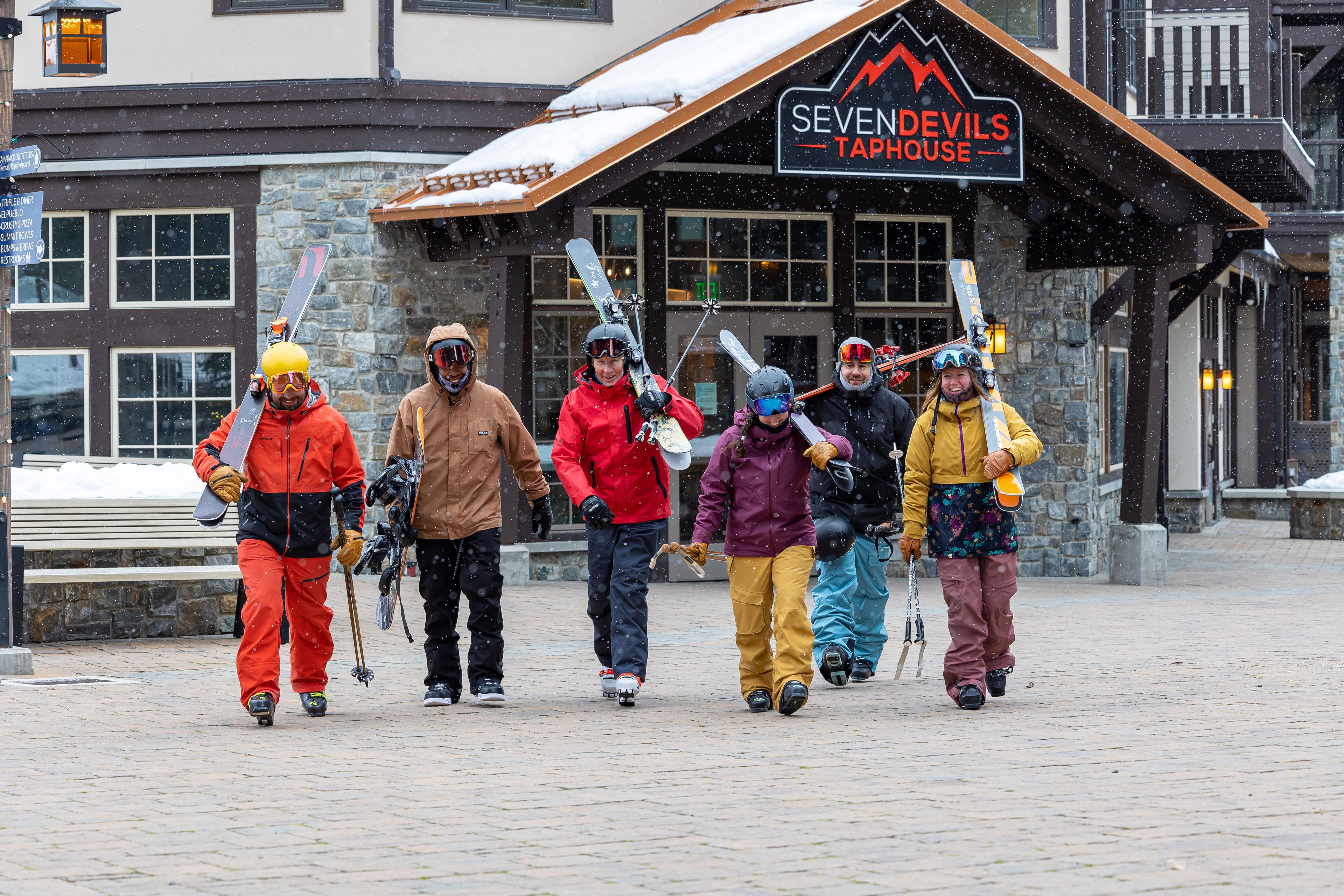 A group of people with skis and ski poles walking in front of a building with a sign that reads 'SEVEN DEVILS TAPHOUSE'.