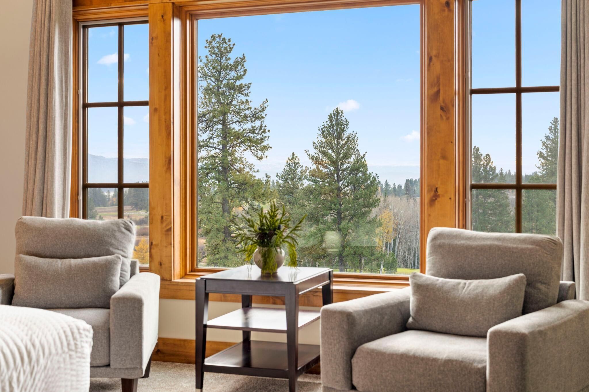 A room with two grey armchairs, a small table with a plant, and large windows offering a view of trees and mountains.