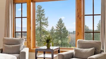 A room with two grey armchairs, a small table with a plant, and large windows offering a view of trees and mountains.