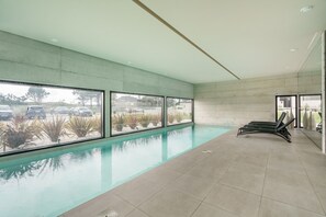 Ein moderner, überdachter Pool mit großen Glaswänden, Fliesenboden und einer Liegebereich mit Liegestühlen.