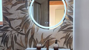 Une salle de bain avec un meuble-lavabo blanc, un miroir rond avec un éclairage intégré et un motif floral sur le mur.