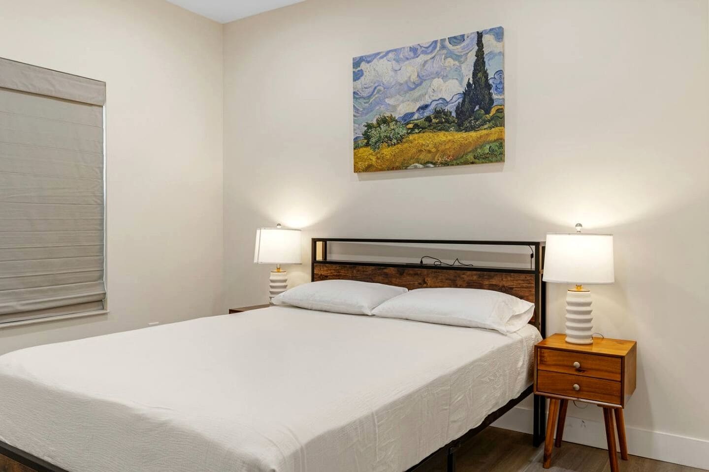 Une chambre à coucher avec un lit, deux tables de chevet avec des lampes, un tableau au mur et une fenêtre avec des stores.