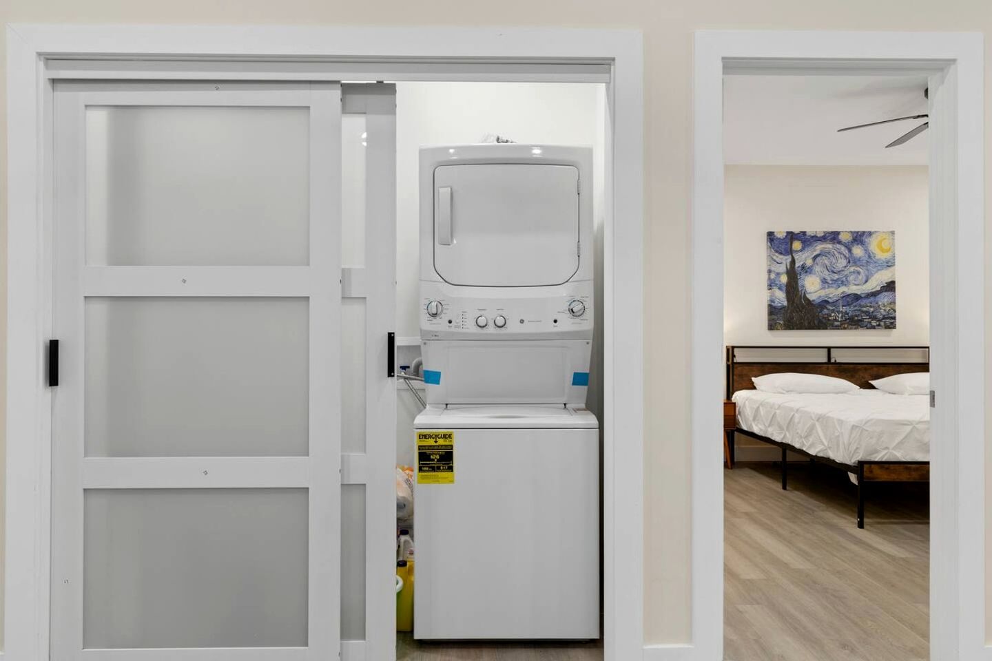 A laundry room with a washing machine and dryer, a bed with white bedding, and a painting on the wall.