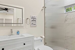 Ein Badezimmer mit Waschbecken, Spiegel und Toilette. Es gibt einen Deckenventilator, ein Hinweis zu empfindlichen Rohren und eine Dusche mit Glastür.