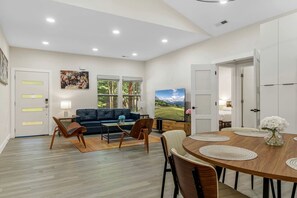 Ein modernes Wohnzimmer mit einer Couch, einem Couchtisch und Sesseln, ein Essbereich mit Tisch und Stühlen sowie ein großes Fernsehgerät.