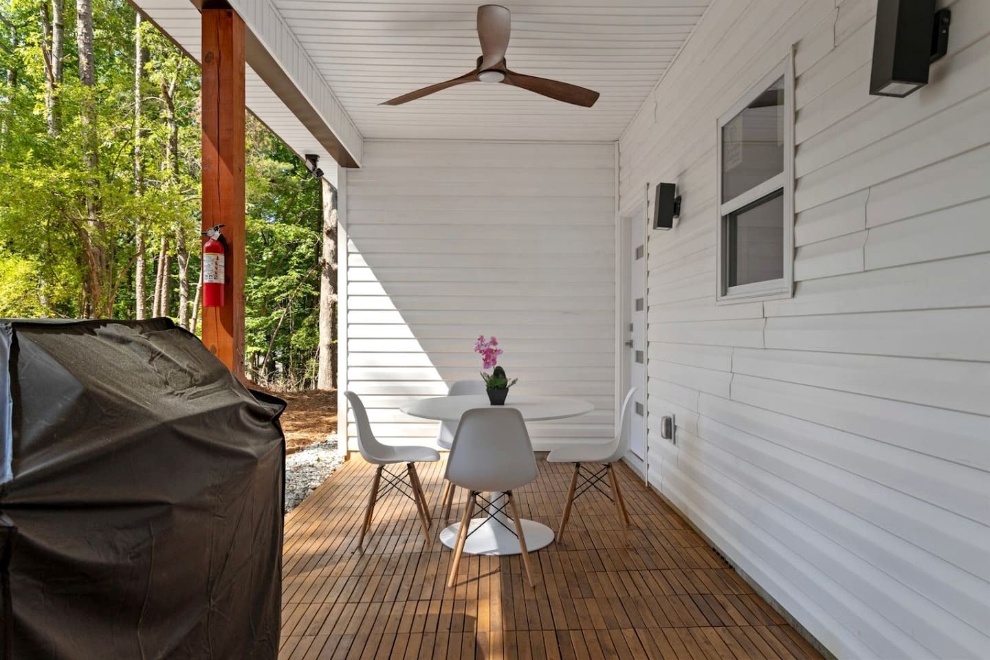 A covered outdoor area with a ceiling fan, two white chairs, a small table with a potted plant, and a barbecue grill.