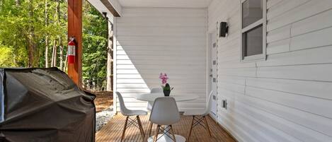 A covered outdoor area with a ceiling fan, two white chairs, a small table with a potted plant, and a barbecue grill.