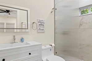 Ein Badezimmer mit Waschbecken, Spiegel und Toilette. Es gibt einen Deckenventilator, ein Hinweis zu empfindlichen Rohren und eine Dusche mit Glastür.