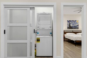 Une buanderie équipée d’un lave-linge et d’un sèche-linge, un lit avec du linge de lit blanc et un tableau accroché au mur.