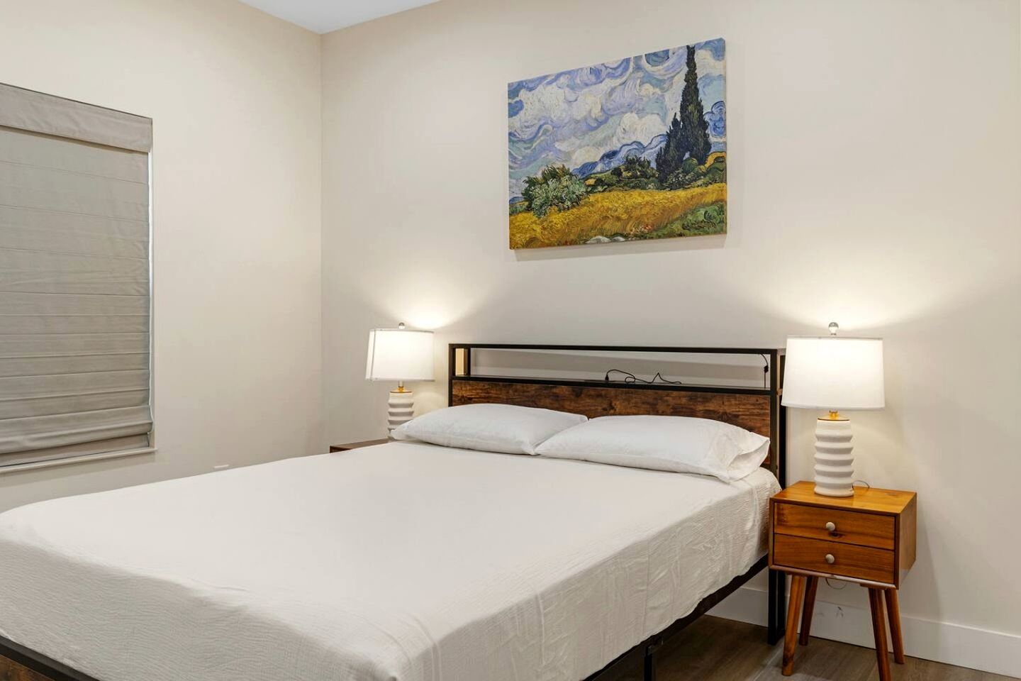Un dormitorio con cama, dos mesitas de noche con lámparas, un cuadro en la pared y una ventana con persianas.