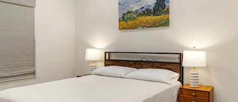 Un dormitorio con cama, dos mesitas de noche con lámparas, un cuadro en la pared y una ventana con persianas.