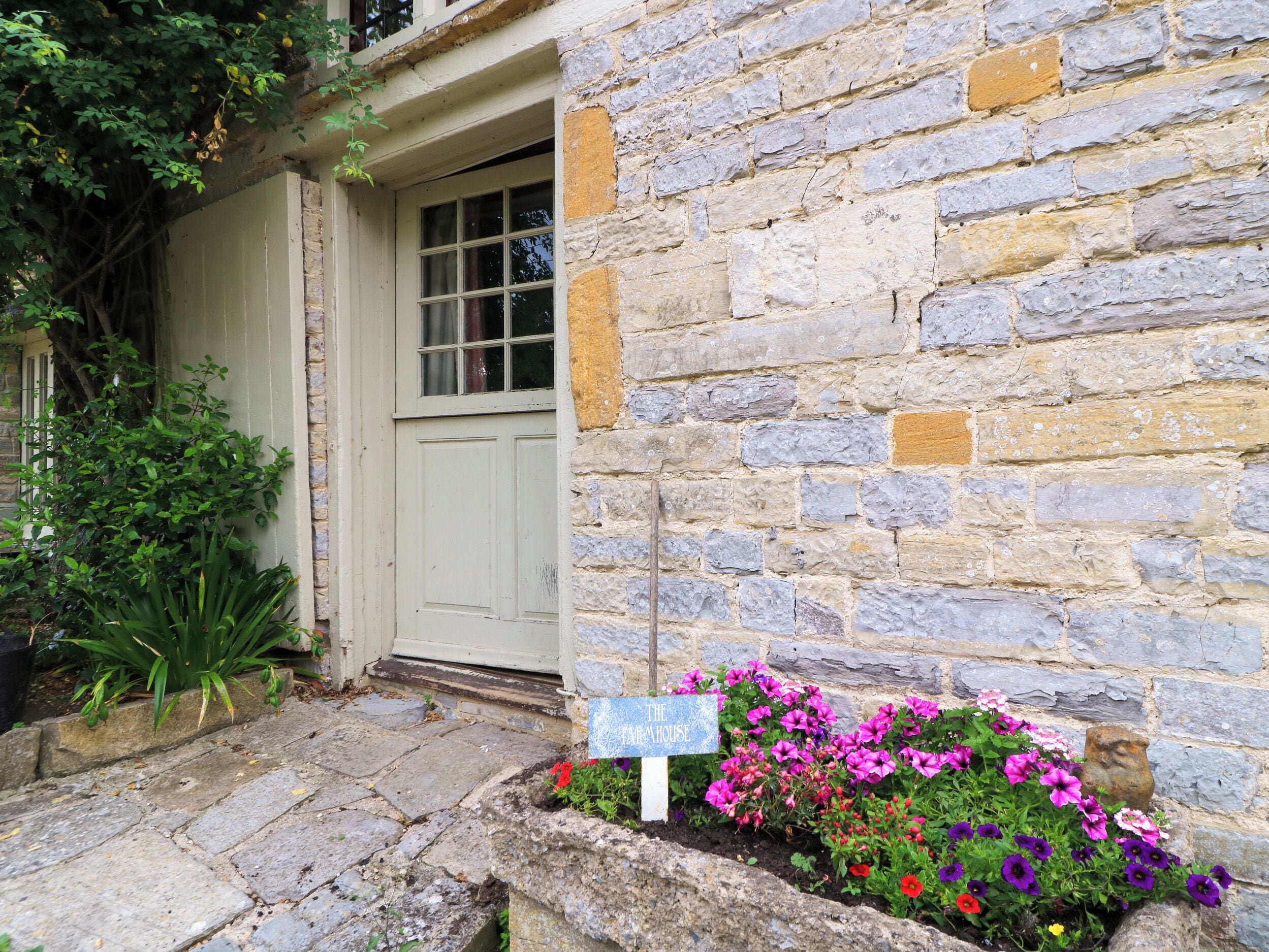 A stone building with a white door, a sign reading 'The House,' and a flower bed with various flowers.