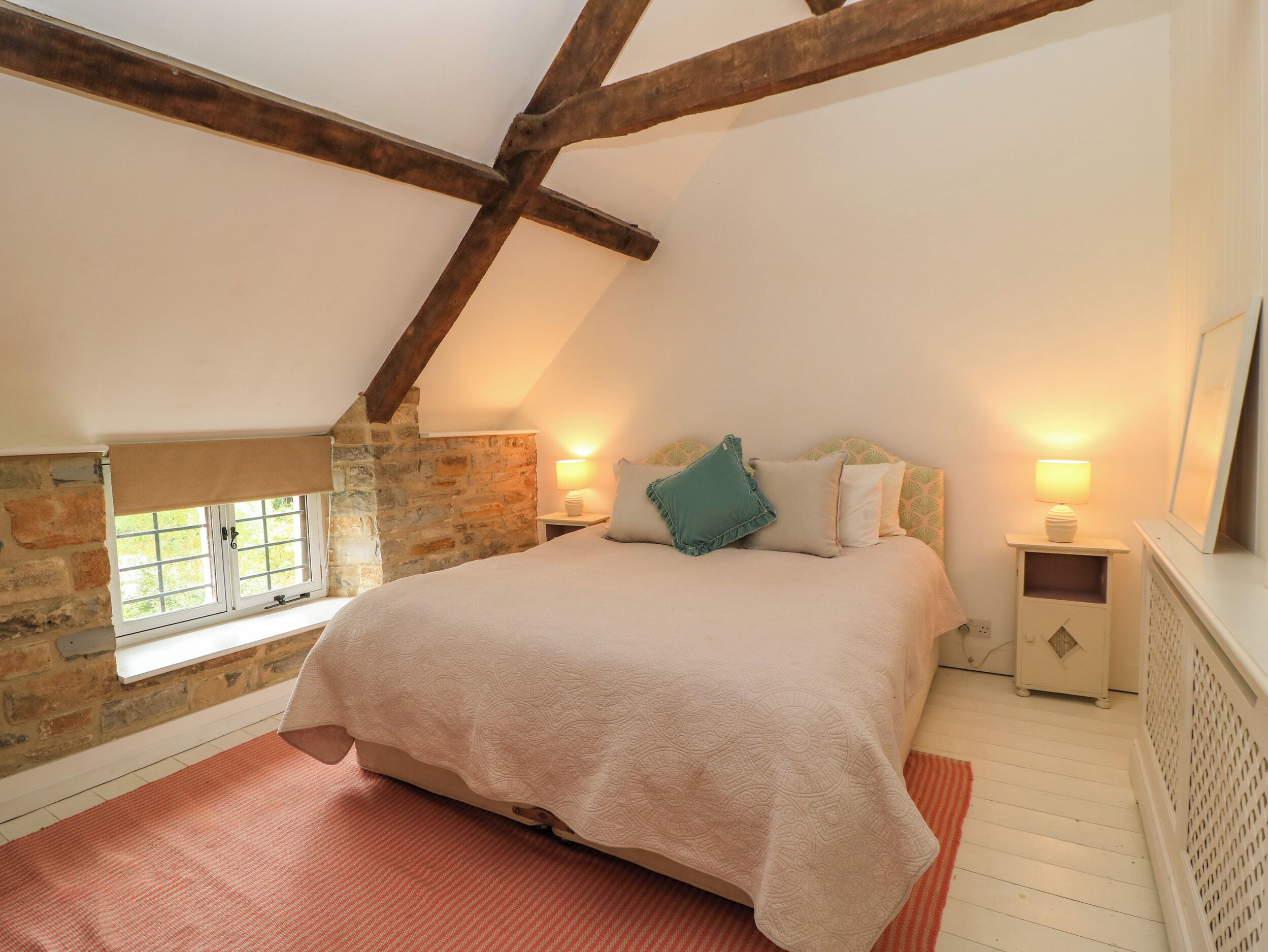 Ein Schlafzimmer mit schrägen Wänden, einer Steinwand, einem Bett mit gemustertem Überwurf, zwei Nachttischlampen und einem Fenster mit Ausblick.