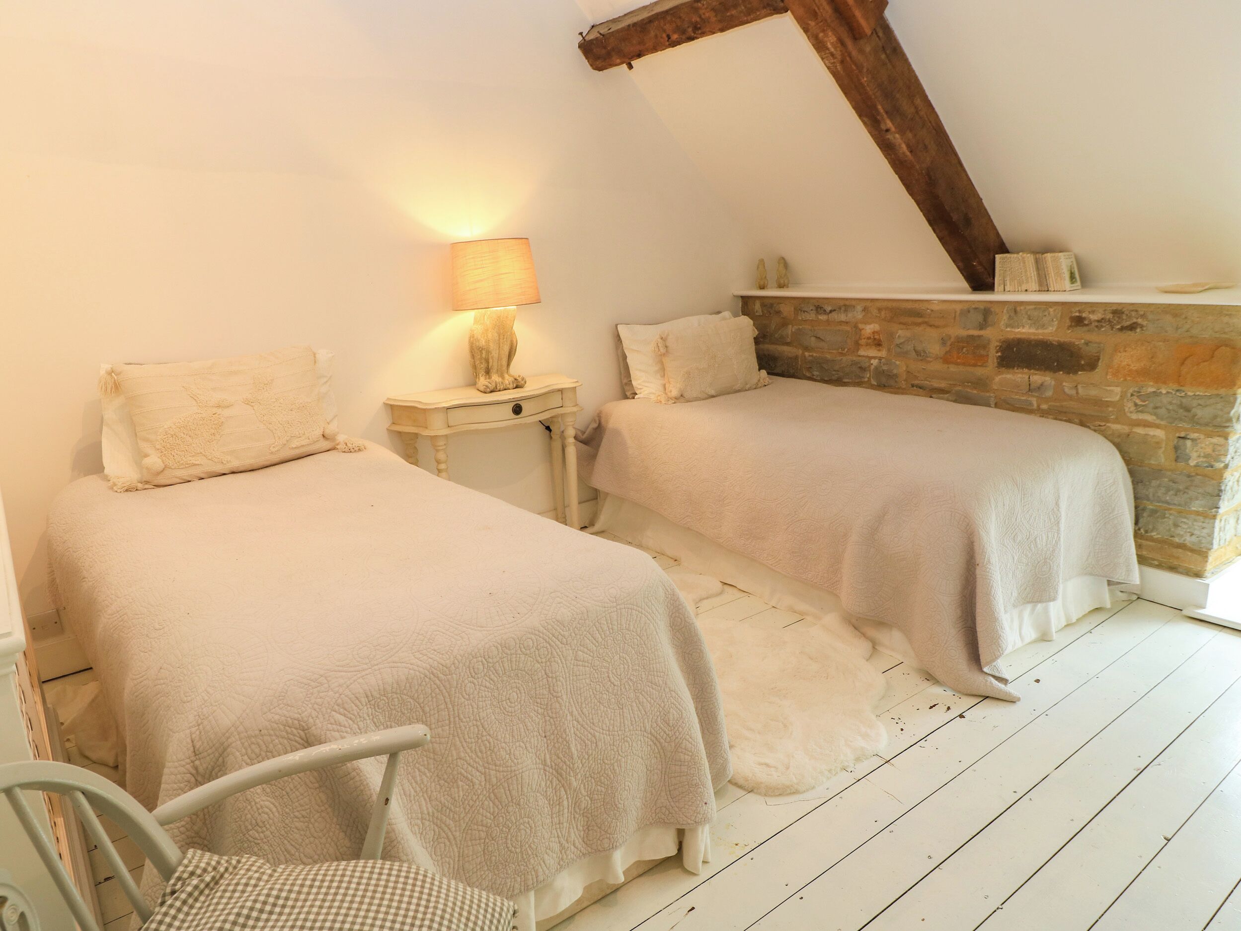 A bedroom with two beds, a nightstand with a lamp, a chair, and a stone wall.