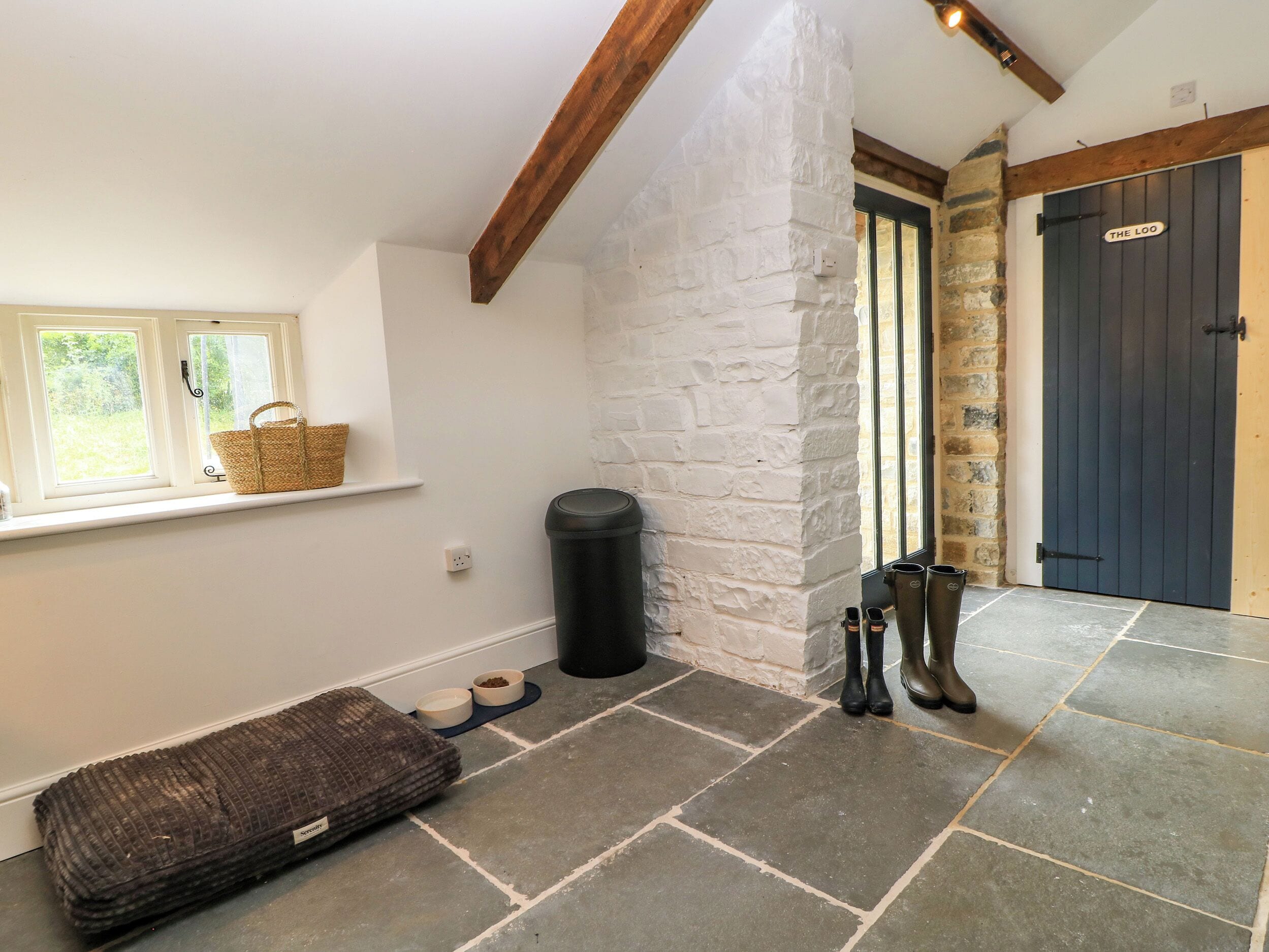 A room with a stone wall, a window with a basket, a black trash bin, a pet bed, pet bowls, and a door labeled 'THE LOO'.