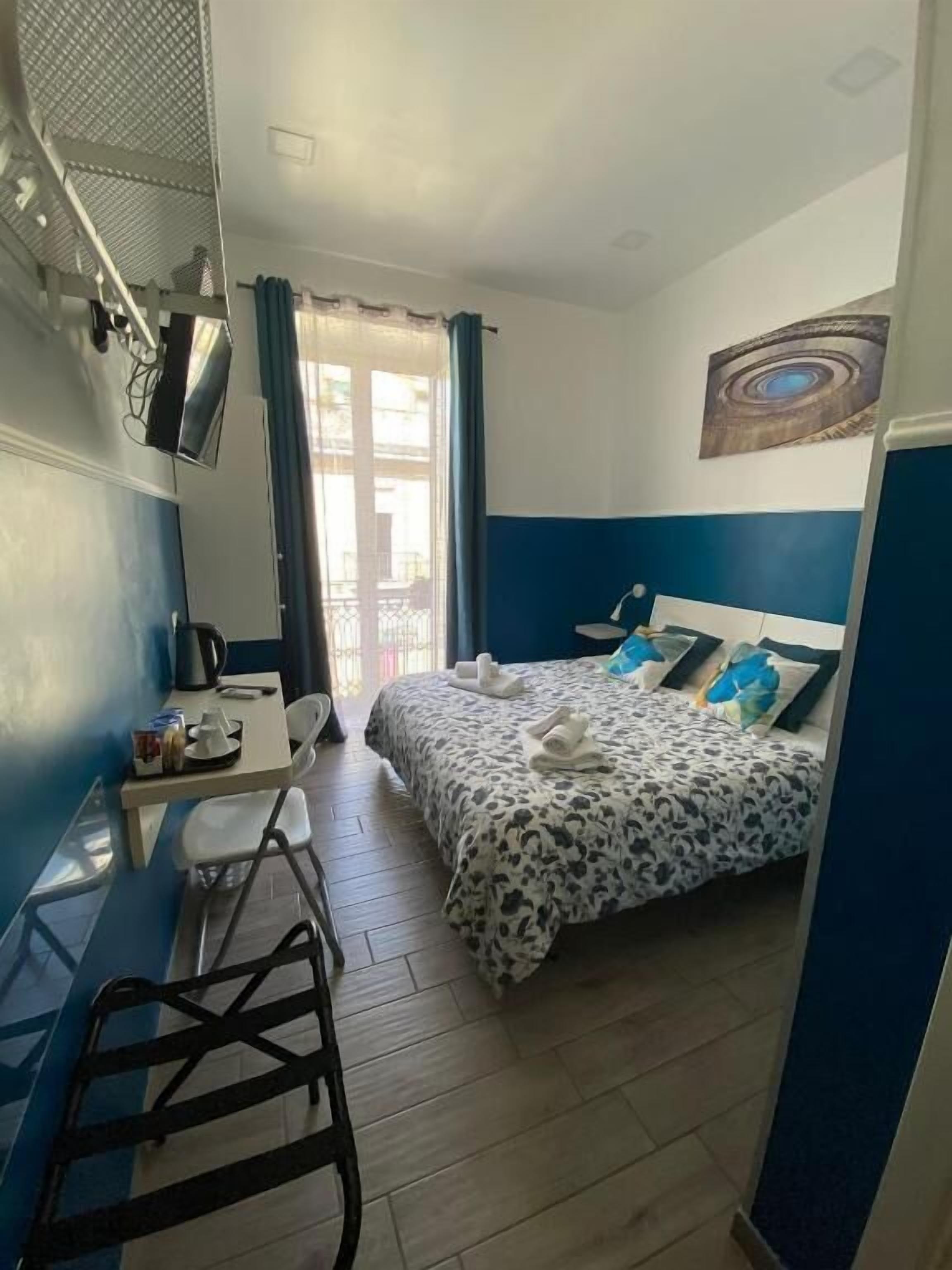 Una habitación pequeña y funcional con cama, escritorio y silla. Tiene una ventana con cortinas, un televisor de pared y un cuadro sobre la cama.