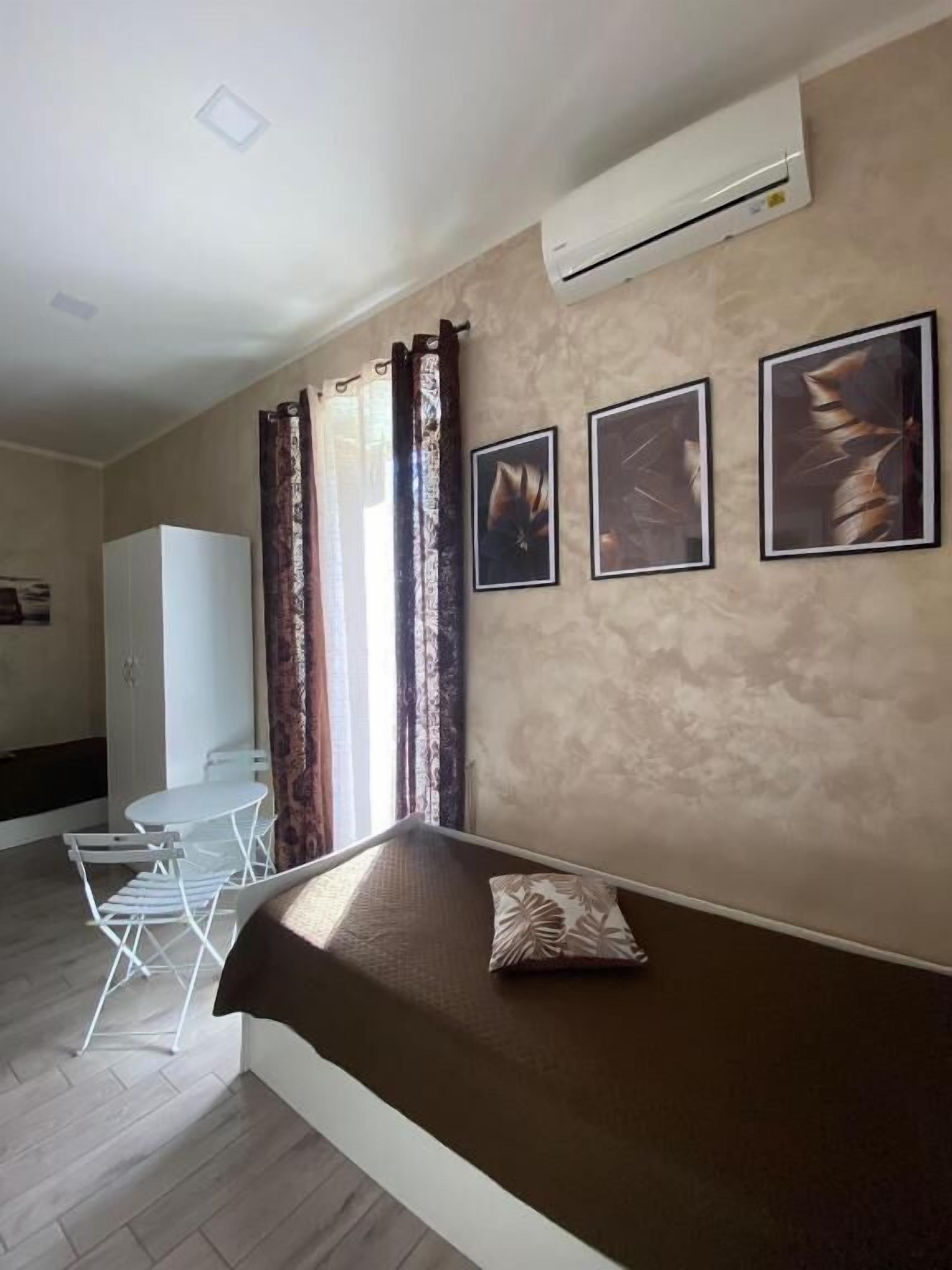 A room with a bed, a white wardrobe, a dining table with chairs, and a window with curtains.