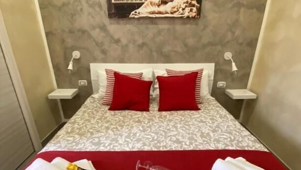 A hotel room with a bed, red pillows, a red bedspread, a painting on the wall, and towels on the bed.