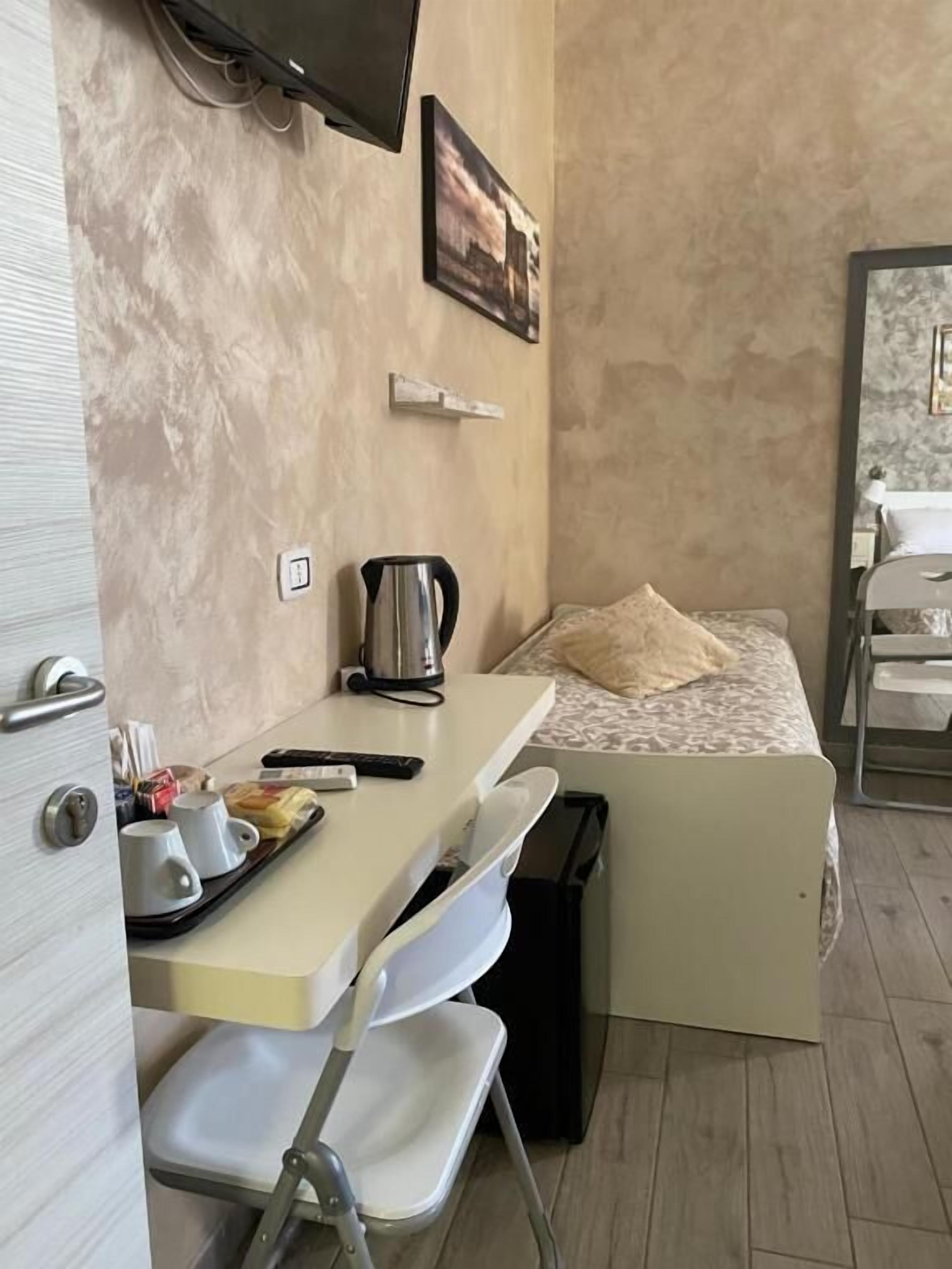 Ein kleines Zimmer mit einem Bett, einem kleinen Tisch mit Wasserkocher, einem Sessel und einem an der Wand befestigten Fernseher.