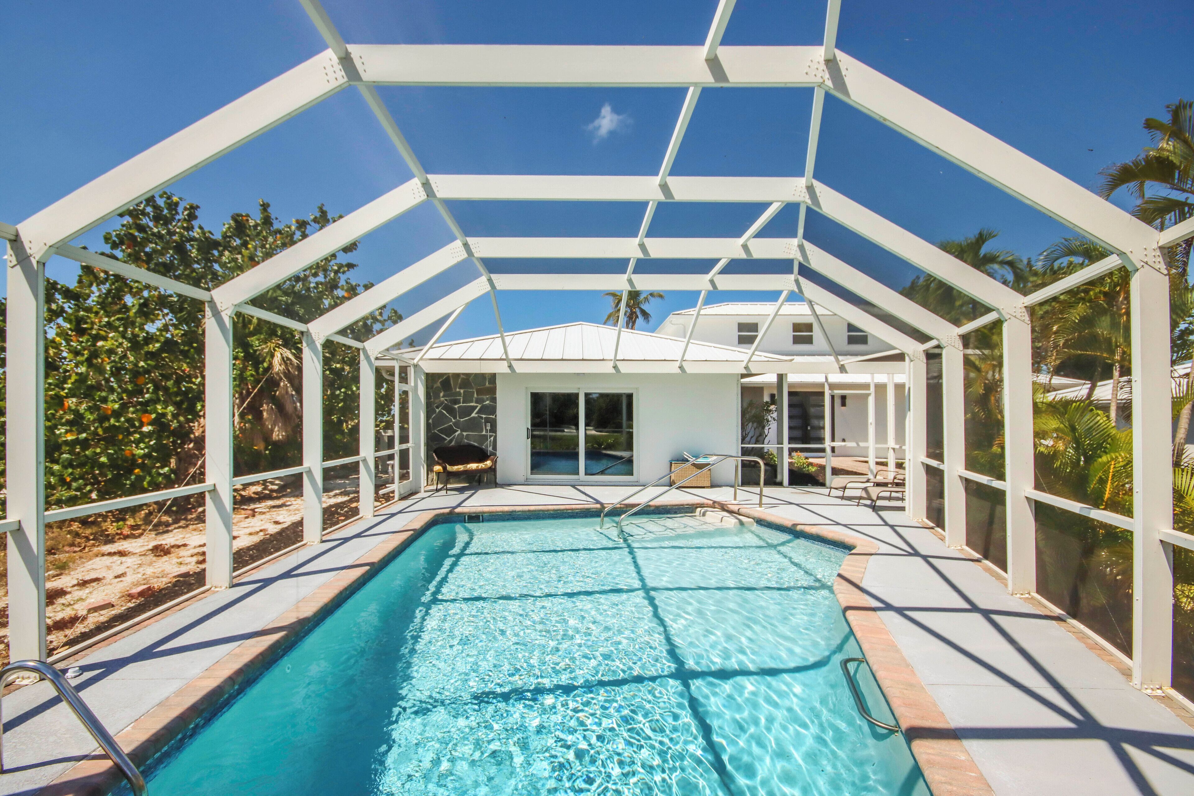 A covered swimming pool with a glass enclosure, a lounge area with chairs, and a view of palm trees.