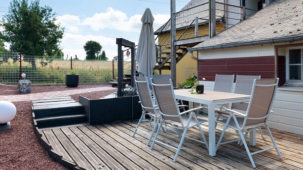 A deck with a table and chairs, a pergola with an umbrella, and a building in the background.